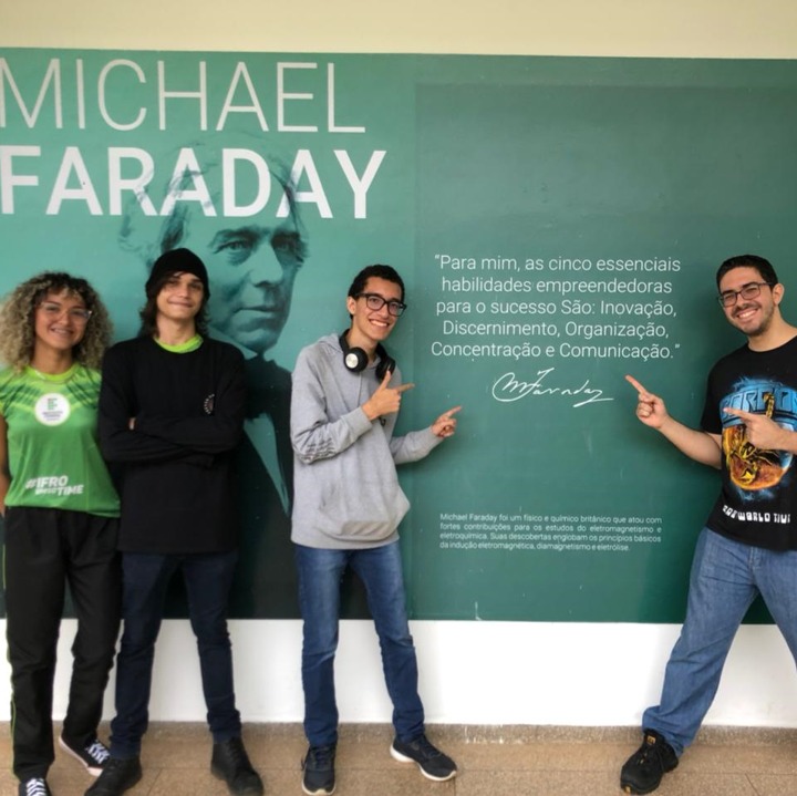 Os alunos Amanda Guilherme e Carlos Henrique posam junto ao professor José Diogo Luna em frente ao painel de Michael Faraday